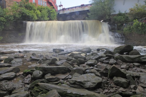 A waterfall cascades over rocks, with a bridge visible above and lush greenery surrounding the scene.