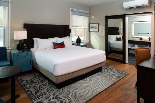 A cozy hotel room featuring a queen bed, wooden furniture, a large mirror, and a colorful area rug.