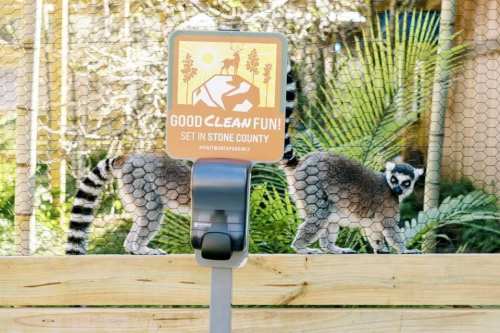 A sign reading "Good Clean Fun! Set in Stone County" in front of two lemurs behind a wire fence.
