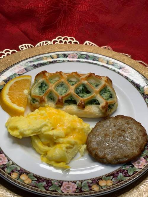A plate with a spinach pastry, scrambled eggs with cheese, a sausage patty, and a slice of orange on a decorative plate.
