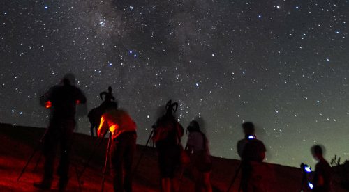 A group of people with tripods observing the starry night sky, illuminated by faint red lights.
