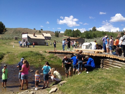 A lively outdoor scene with people gathered by a stream, exploring, and enjoying a sunny day near historic buildings.