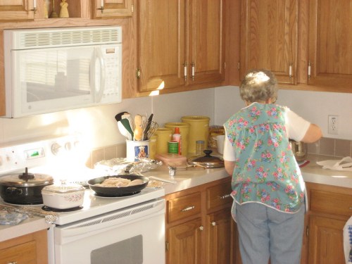 An elderly woman in a floral apron prepares food in a sunny kitchen with wooden cabinets and various cooking utensils.