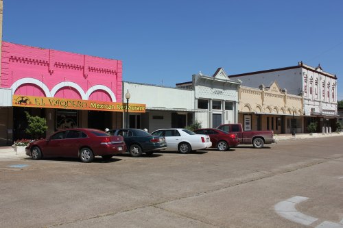 A row of buildings in a small town, featuring a pink Mexican restaurant and parked cars along the street.
