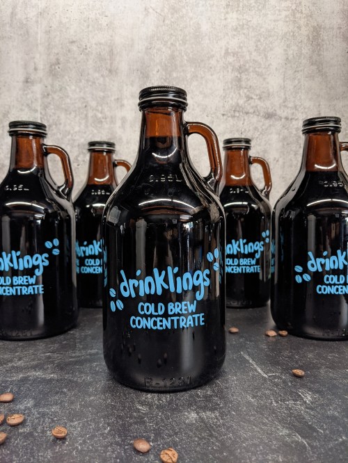 A close-up of a dark glass bottle labeled "drinklings Cold Brew Concentrate" with several bottles in the background.