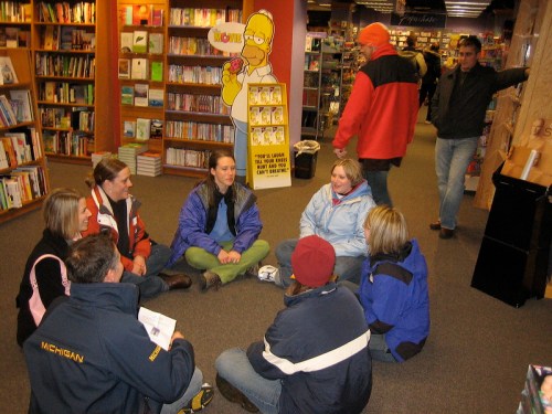 A group of people sitting on the floor in a bookstore, chatting, with a cartoon character display in the background.
