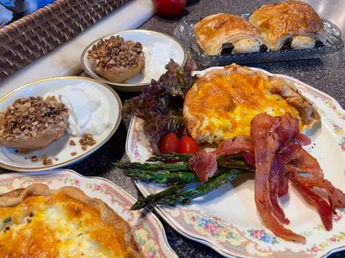 A colorful breakfast spread featuring quiche, bacon, asparagus, yogurt with granola, and pastries on decorative plates.