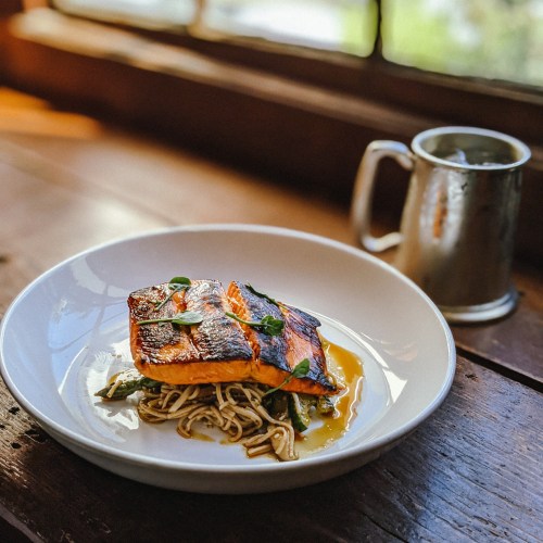 A plate of grilled salmon on a bed of noodles, garnished with greens, beside a metal mug on a wooden table.