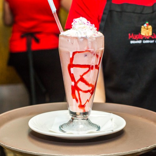 A tall glass of strawberry milkshake topped with whipped cream and a drizzle of strawberry sauce, served on a plate.