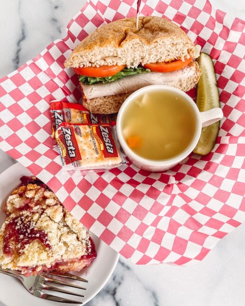 A sandwich with turkey and veggies, a cup of soup, a pickle, crackers, and a slice of berry pie on a checkered tablecloth.