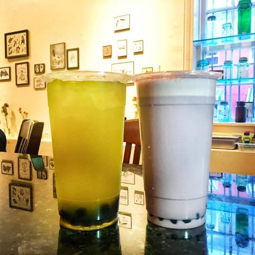 Two drinks on a table: one yellow with bubbles and one light purple with tapioca pearls, in a cozy café setting.