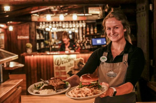 A smiling server holds two plates of food, featuring a steak and a burger with fries, in a cozy restaurant setting.