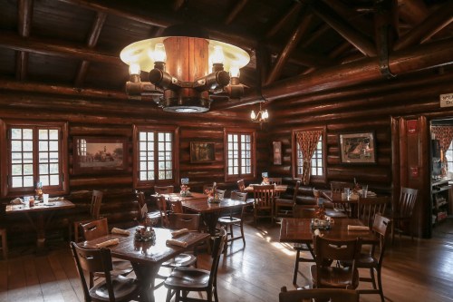 Cozy log cabin interior with wooden tables, chairs, and warm lighting, featuring rustic decor and large windows.
