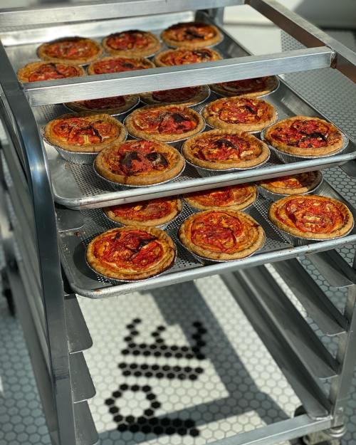 A metal cart filled with freshly baked mini pizzas, topped with tomatoes, on a tiled floor.