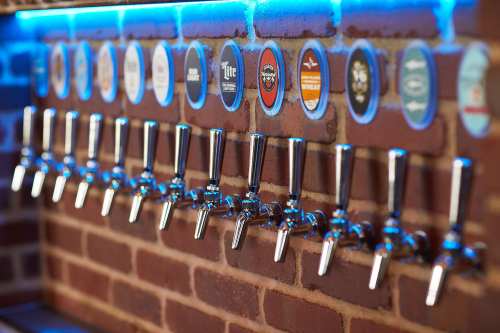 A row of shiny beer taps against a brick wall, with various beer logos displayed above.