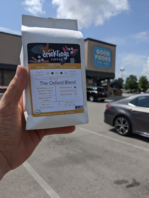 A hand holds a bag of "The Oxford Blend" coffee outside a store named Good Foods.
