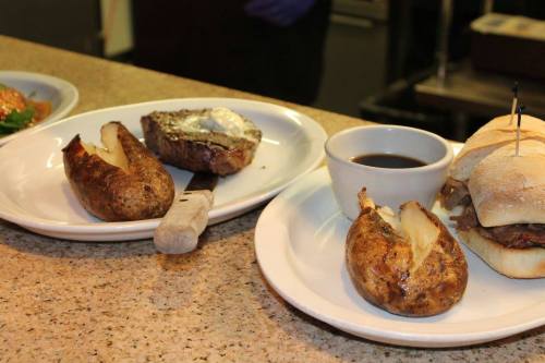 A plate with two baked potatoes, a small bowl of sauce, and a sandwich on a white plate, set on a countertop.
