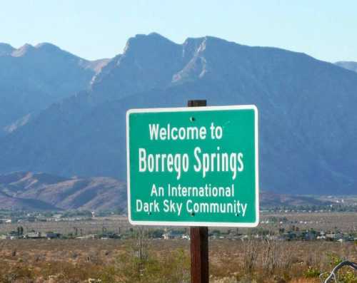 Green sign reading "Welcome to Borrego Springs, An International Dark Sky Community" with mountains in the background.