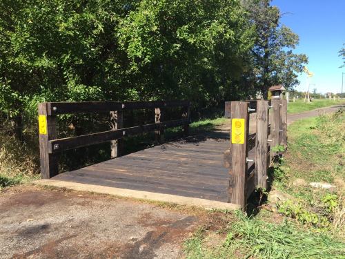 A wooden bridge over a path, surrounded by greenery and trees under a clear blue sky.