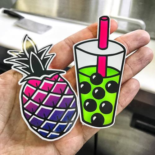 A hand holding two colorful stickers: a pineapple and a drink with green liquid and black bubbles.