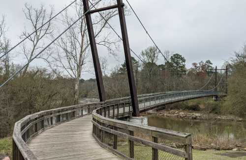 A wooden walkway leads to a suspension bridge over a river, surrounded by trees and a cloudy sky.