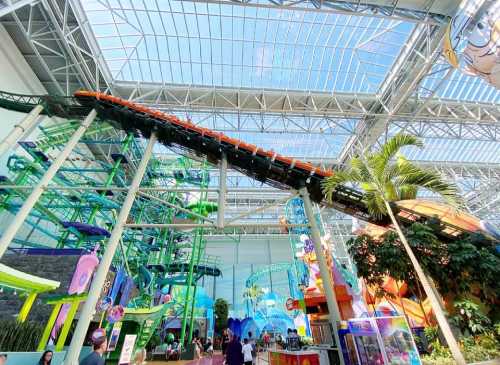 Indoor amusement park featuring a roller coaster, vibrant decorations, and lush greenery under a glass ceiling.