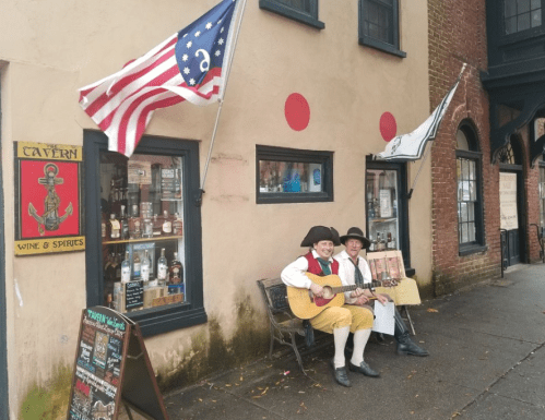 Two musicians in colonial attire play guitar outside a historic building with flags and a sign for a wine shop.