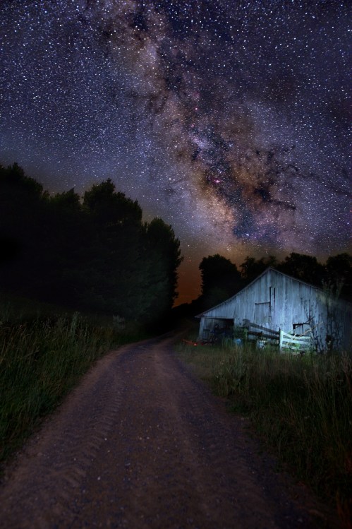 A gravel path leads to a rustic barn under a starry night sky filled with the Milky Way. Trees frame the scene.