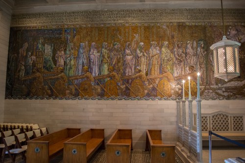 Mural depicting historical figures and scenes, with wooden benches and candle holders in a well-lit interior space.