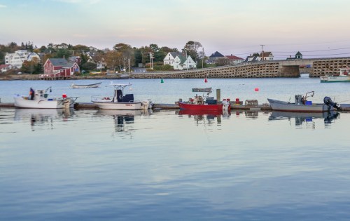 A calm harbor scene with several boats docked, surrounded by quaint houses and a bridge in the background.