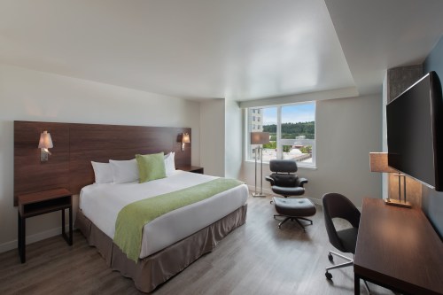 A modern hotel room featuring a king bed, desk, chair, and large window with natural light.