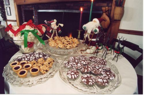 A festive table displays an assortment of holiday cookies and treats, with a cozy fireplace in the background.