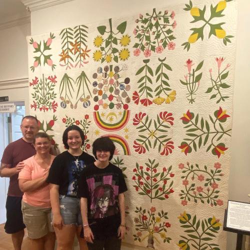 A group of four people stands in front of a colorful floral quilt displayed on a wall.