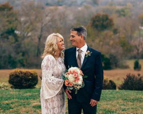 A couple smiles at each other, holding a bouquet, with a scenic outdoor background. They appear joyful and in love.