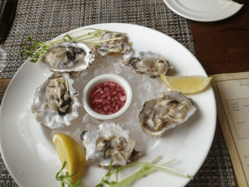 A plate of fresh oysters on ice, garnished with lemon wedges, microgreens, and a small bowl of red onion relish.