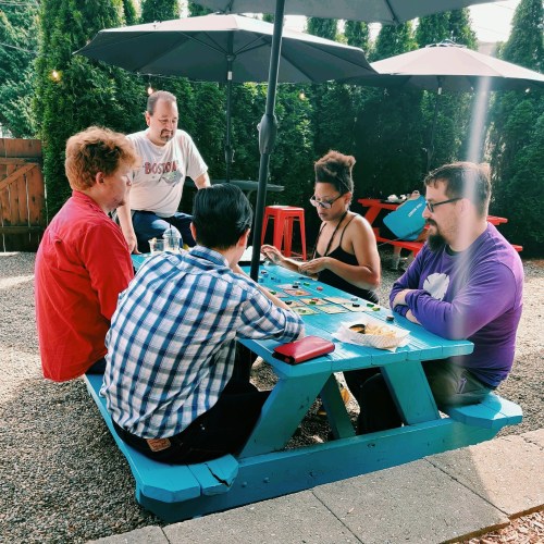 A group of five people sitting around a blue picnic table, playing a game outdoors under umbrellas.