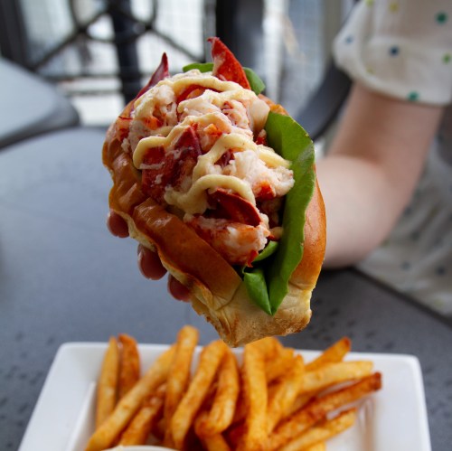 A hand holding a lobster roll with lettuce and mayo, accompanied by a plate of golden fries.