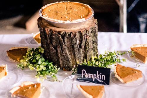 A rustic display featuring a pumpkin pie on a wooden stump, surrounded by slices and greenery.