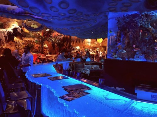 A vibrant bar scene with an illuminated blue countertop, surrounded by a lively restaurant featuring ocean-themed decor.
