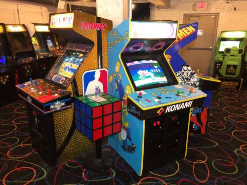 Two vintage arcade machines, one featuring "X-Men" and the other "The Simpsons," alongside a Rubik's Cube display.