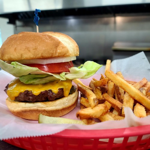 A cheeseburger with lettuce, tomato, and a blue pick on a bun, served with a side of golden French fries.