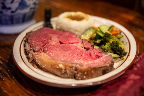 A plate featuring a large slice of prime rib, rice with gravy, and a side of mixed vegetables including broccoli and zucchini.
