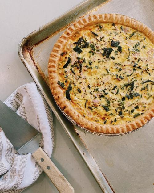 A freshly baked quiche with a golden crust, sitting on a baking tray next to a striped kitchen towel and a spatula.