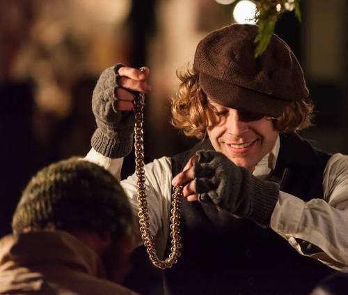 A young man in vintage attire holds a gold chain, smiling as he interacts with another person.