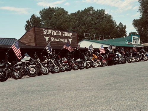 A row of motorcycles parked outside Buffalo Jump Steakhouse, adorned with American flags. Trees and a blue sky in the background.