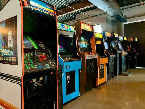 A row of classic arcade game machines in a well-lit space, featuring colorful screens and retro designs.