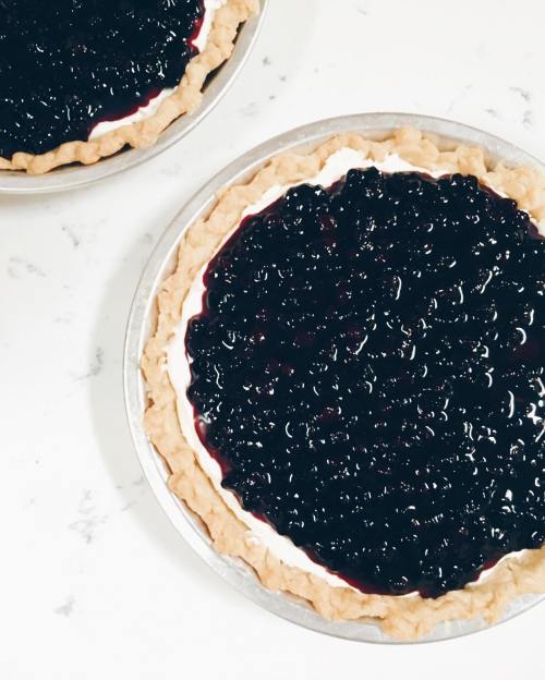 Two blueberry pies with a glossy berry topping on a flaky crust, set on a light marble surface.