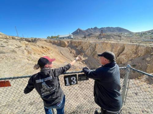 Two people stand by a fence, pointing at a large excavation site with mountains in the background under a clear blue sky.