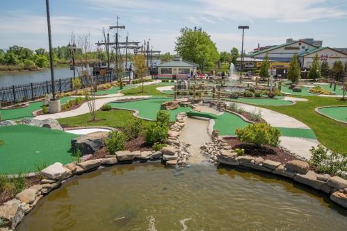 A scenic mini-golf course with green turf, water features, and a view of a river and nearby buildings.