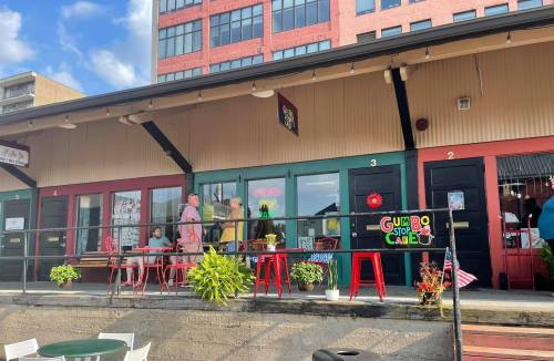 A colorful storefront with outdoor seating, featuring a sign for "Gumbo Stop Café" and people walking by.